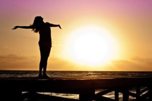 The picture of a woman standing on a pier holding her hands up to the sunset.