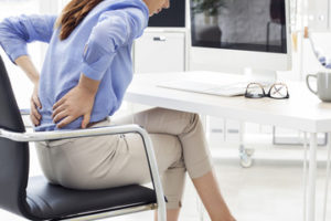 The picture of a woman sitting in a chair, holding her back, as she is in severe pain.
